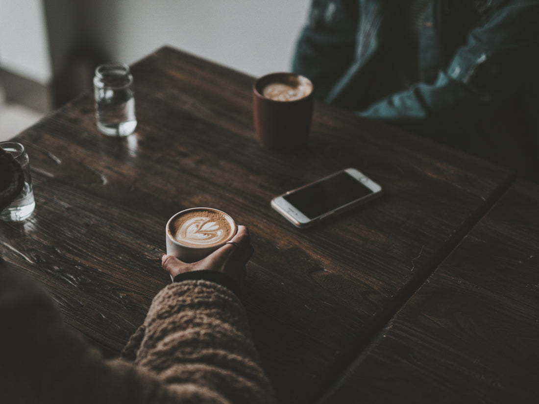 A person holding a cup of latte with heart-shaped latte art at a wooden café table, beside a phone and another coffee cup — capturing the calm ritual of sharing coffee and noticing each cup’s subtle differences.
