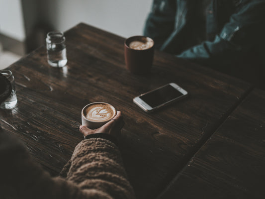 A person holding a cup of latte with heart-shaped latte art at a wooden café table, beside a phone and another coffee cup — capturing the calm ritual of sharing coffee and noticing each cup’s subtle differences.